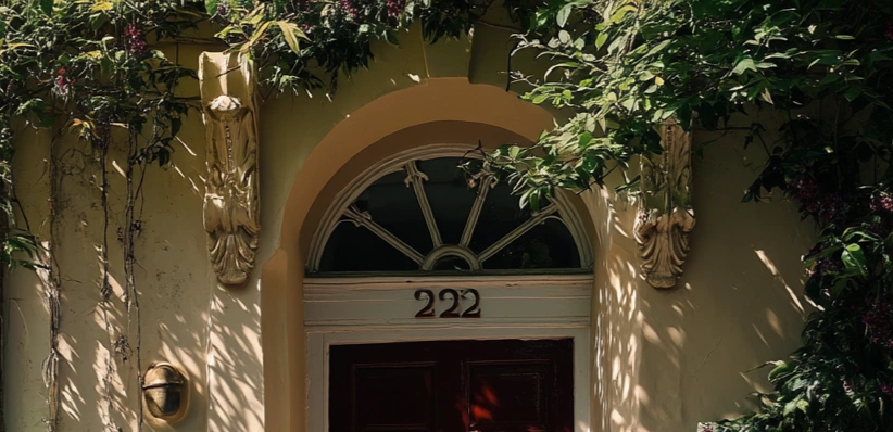 An ornate doorway of a house with the number 222 above a maroon door, framed by stone scrollwork and trailing greenery in warm sunlight