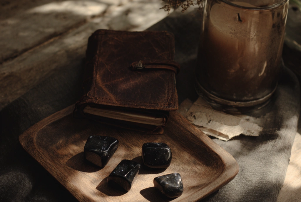 A leather-bound journal on a small wooden tray with four black tumbled stones in front of it, beside a glass-jarred candle and dried herbs on a wooden surface in soft warm light