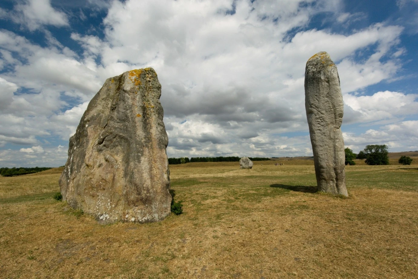 The standing sarsen stones of Avebury henge rising from a green Wiltshire field, with the village of Avebury visible between the stones, photographed in overcast autumn light