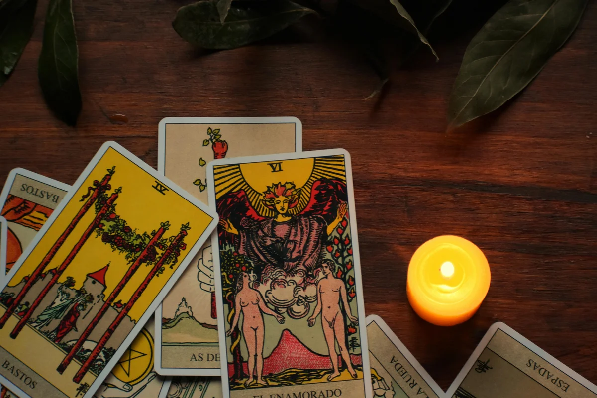 Several Rider-Waite tarot cards spread on a dark wooden surface by candlelight, with The Lovers (VI) prominent in the centre, alongside Wands cards and others labelled in Spanish, surrounded by large green leaves