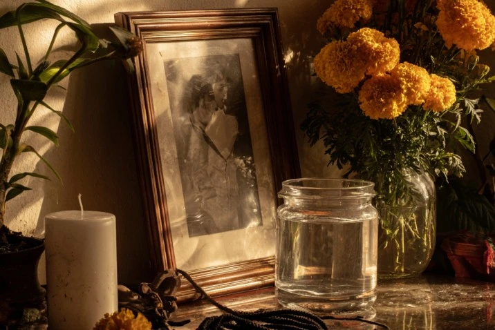 A small ancestor altar with a framed black-and-white photograph, a glass of clean water, fresh marigolds, a single white candle, and a length of black cord, photographed in low evening light