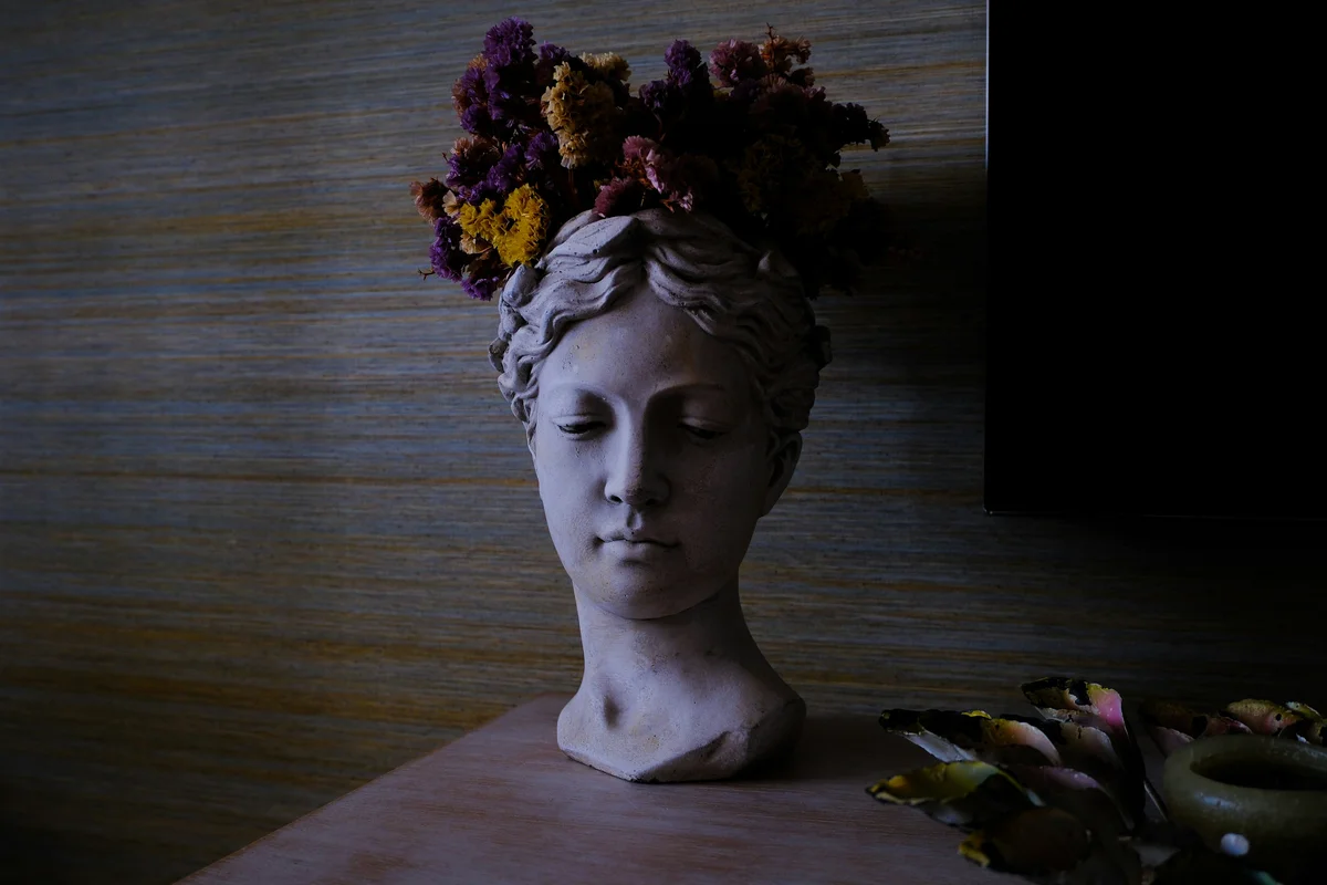 A classical Greek-style plaster bust of a female figure with closed eyes, used as a planter with dried purple, yellow, and rust-coloured flowers crowning the head, resting on a wooden surface beside scattered rose petals in dim moody light