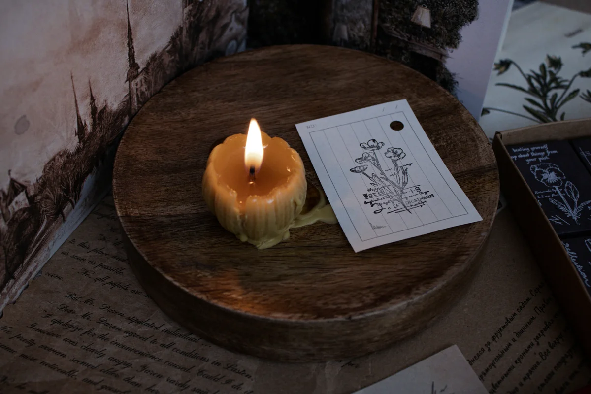 A lit beeswax candle melting onto a round wooden board, placed beside a botanical illustration card, surrounded by aged handwritten letters and botanical print papers in warm candlelight