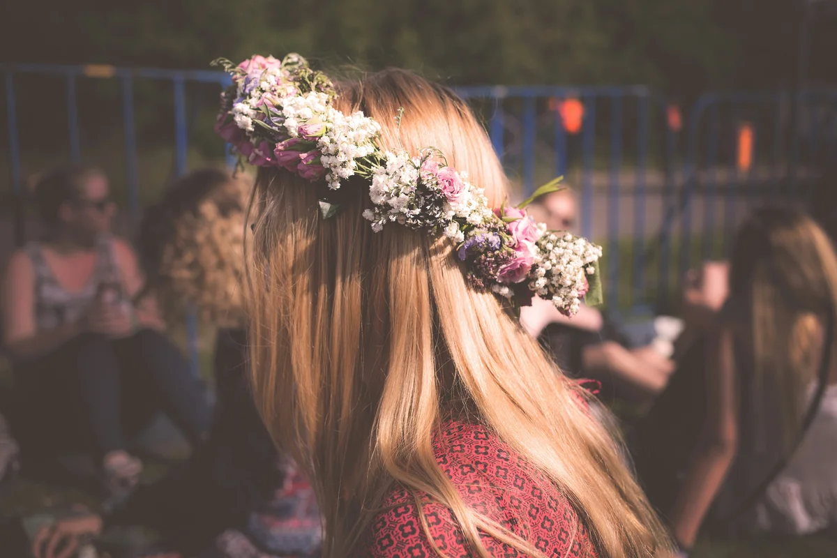 A person with long blonde hair wearing a fresh flower crown of pink roses, white baby's breath, and purple blooms, seen from behind in a crowd at an outdoor gathering in warm evening light