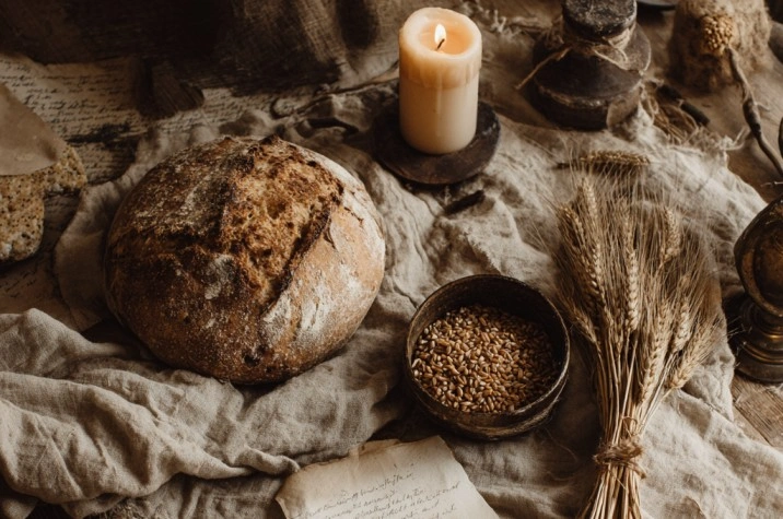 A flat lay of a round handmade loaf cooling on a linen cloth, a small bowl of wheat berries, three ears of dried wheat tied with twine, a beeswax candle, and an open journal with handwritten intentions on a weathered wooden table