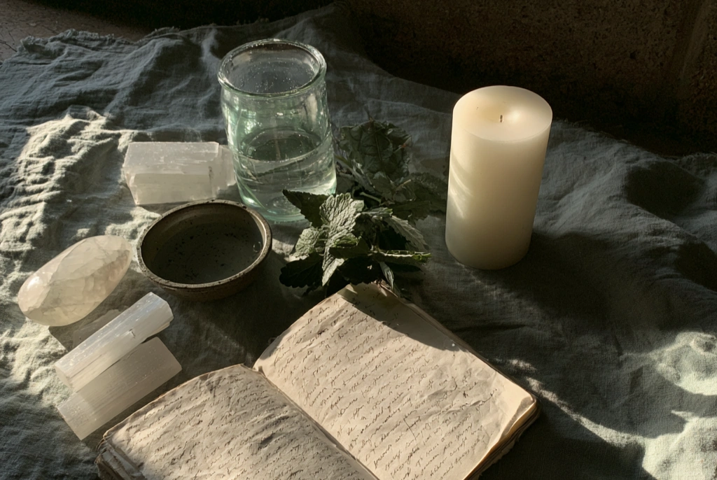 A flat lay of moonstone and selenite, a small bowl of water, fresh lemon balm leaves, an open journal with handwritten reflections, and a single white pillar candle on cool grey linen in soft evening light