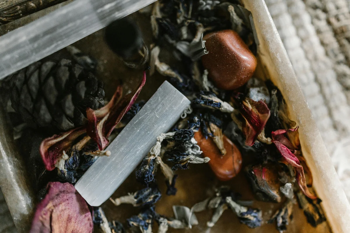 Close-up of a wooden box filled with dried rose petals, dried lavender, polished red jasper tumble stones, a selenite wand, and a pine cone, resting on a woven textile