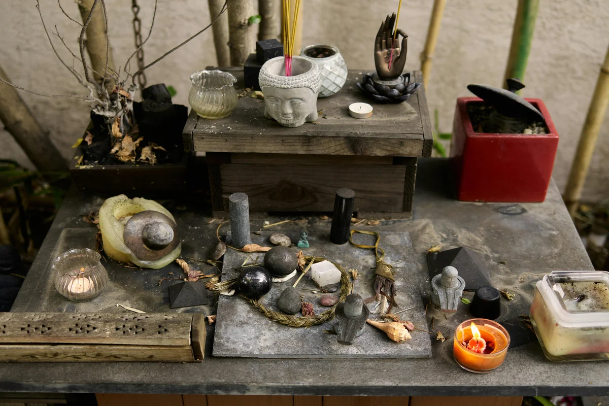 An outdoor altar table spread with a stone Buddha head planter with incense sticks, black obsidian spheres and pyramids, a lit tealight, a selenite wand, a conch shell, dried herbs, river stones, small figurines, and a burning orange pillar candle, set against bare branches and bamboo