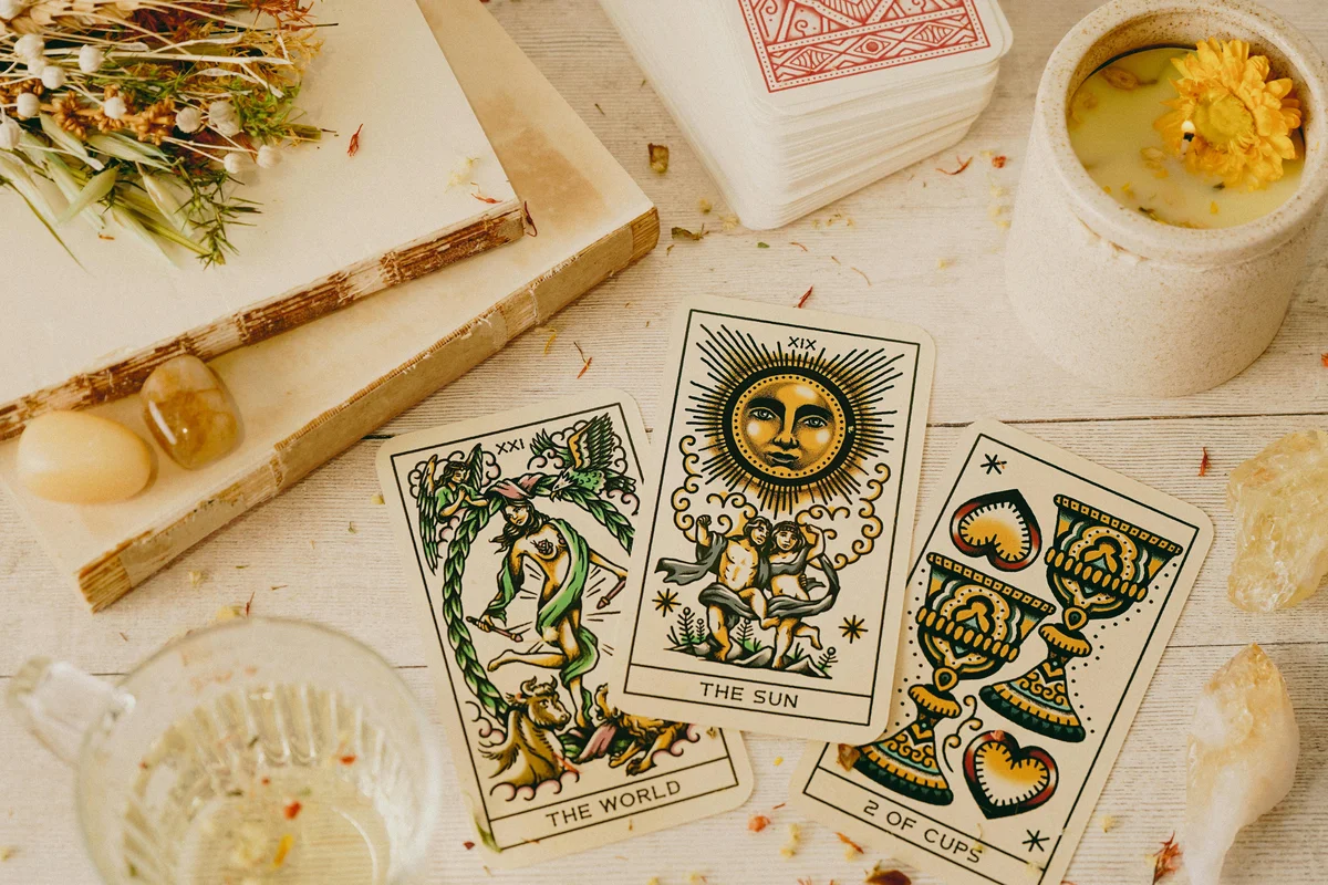 Three tarot cards fanned out on a white wooden surface — The World (XXI), The Sun (XIX), and 2 of Cups — surrounded by scattered dried flower petals, polished crystals, a floral candle, and a stacked tarot deck with a red geometric back design