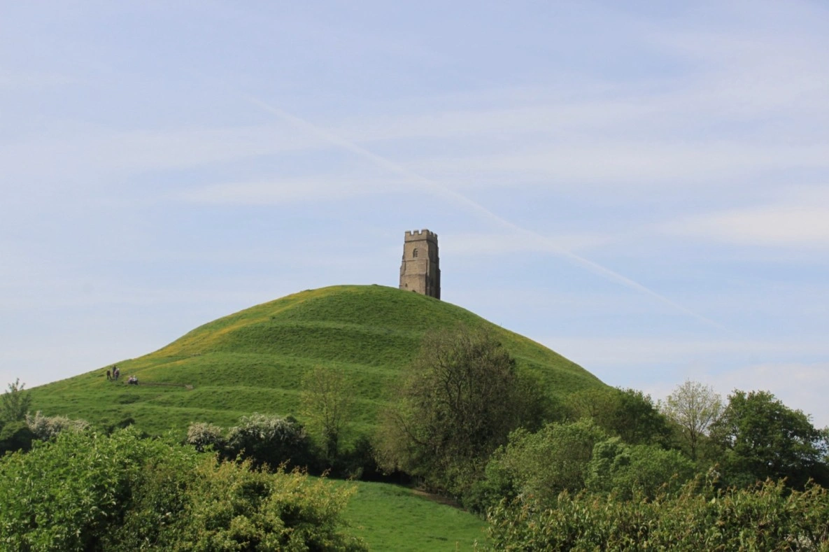The conical silhouette of Glastonbury Tor rising above morning mist in Somerset, with the ruined tower of St Michael's Chapel at its summit against a pale grey sky