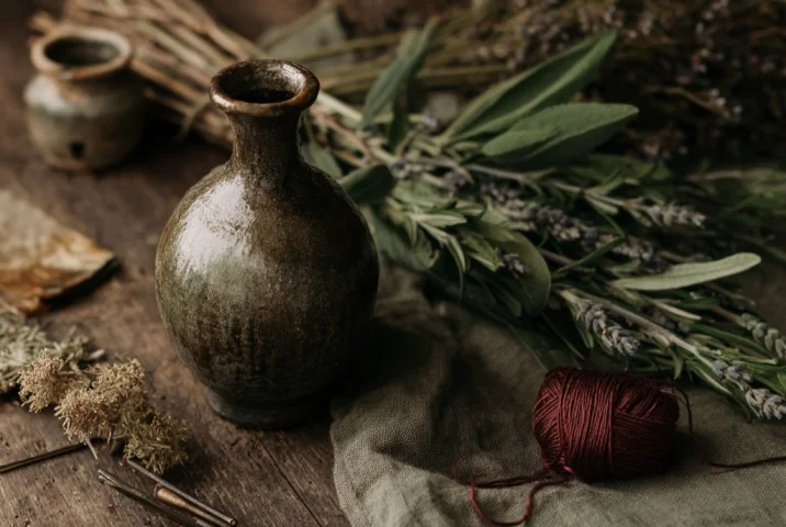 A dark wooden altar surface at Samhain bearing bundles of dried mugwort and rosemary, a handful of apples, a sprig of wormwood, and a black candle, photographed in low warm light