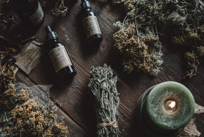 A flat lay of dried herbs, dark glass bottles of condition oil, and a dressed green candle on a wooden surface