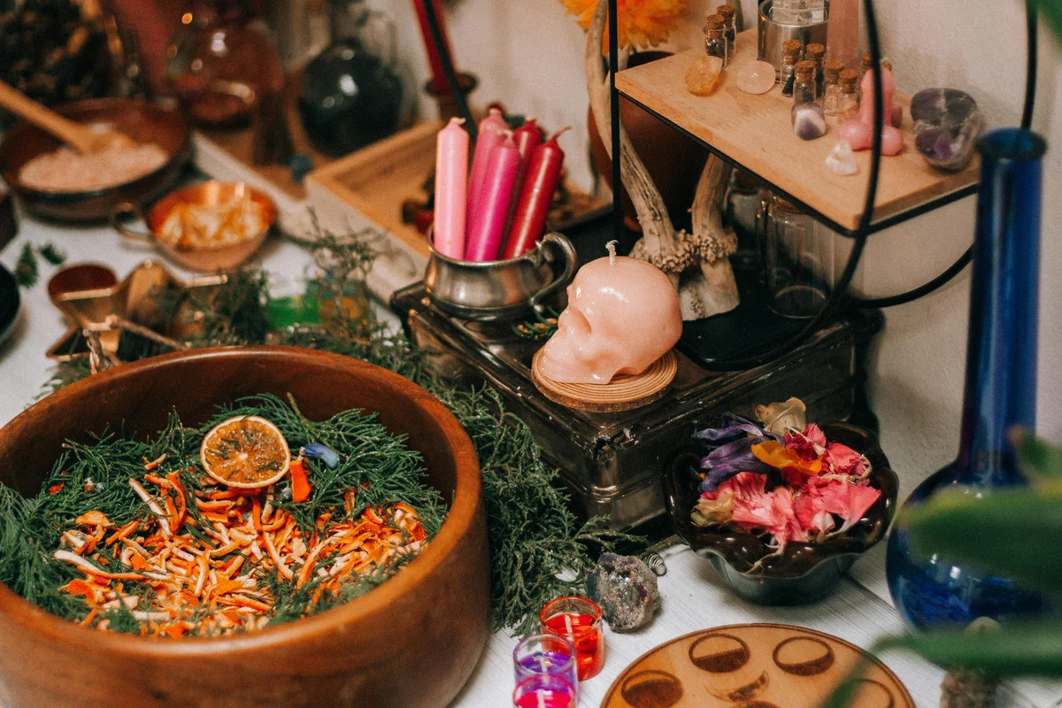 A witch's altar spread with a wooden bowl of dried herbs, orange peel, and greenery, a skull-shaped candle, pink taper candles, small crystal vials, dried flower petals, raw crystals, and coloured spell candles on a white surface