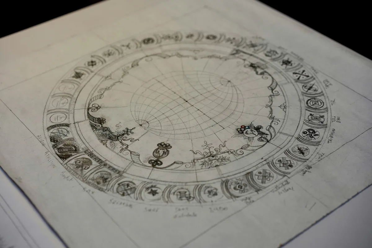 A hand-drawn natal chart wheel on paper, showing the twelve houses, zodiac symbols, and planetary glyphs in pencil, photographed at an angle in moody low light