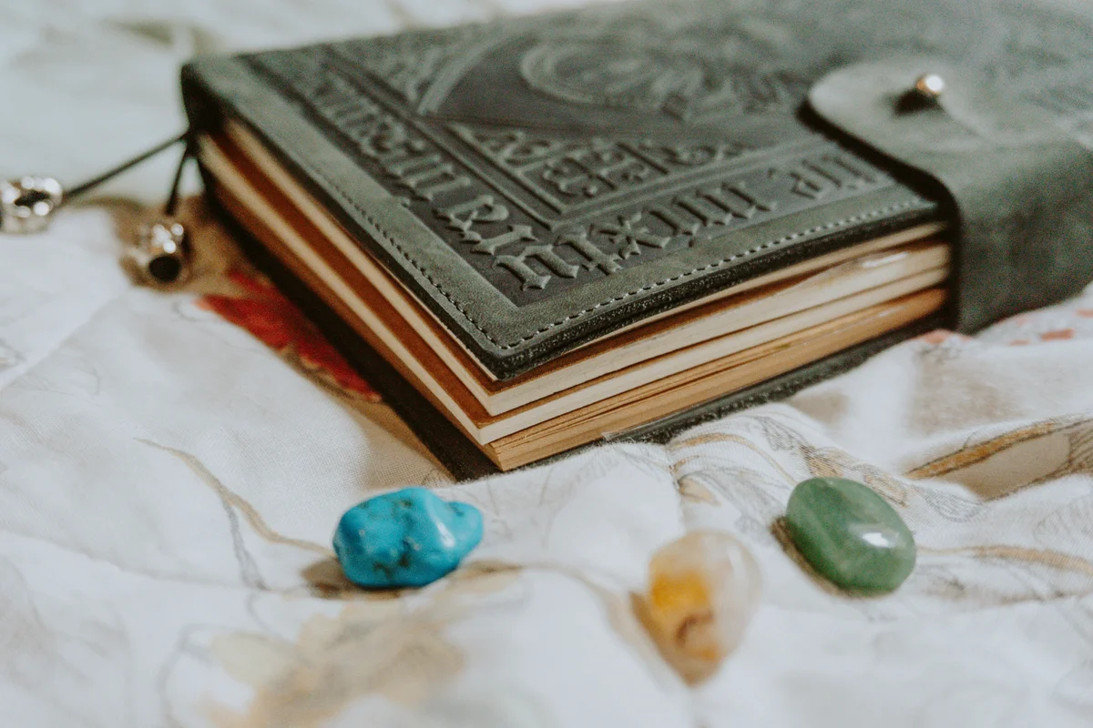 A dark green embossed leather grimoire with Gothic lettering on the cover, lying closed on a white cloth beside three polished crystals — turquoise, citrine, and green aventurine — with a silver charm bracelet nearby