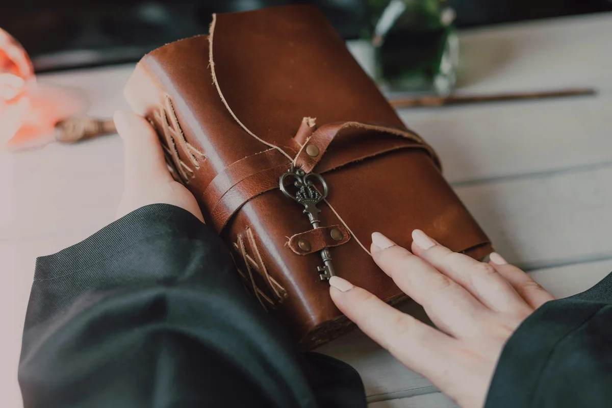 Hands resting on a worn leather-bound journal tied with twine and fastened with an ornate vintage skeleton key, held in the lap of a person wearing a dark green robe