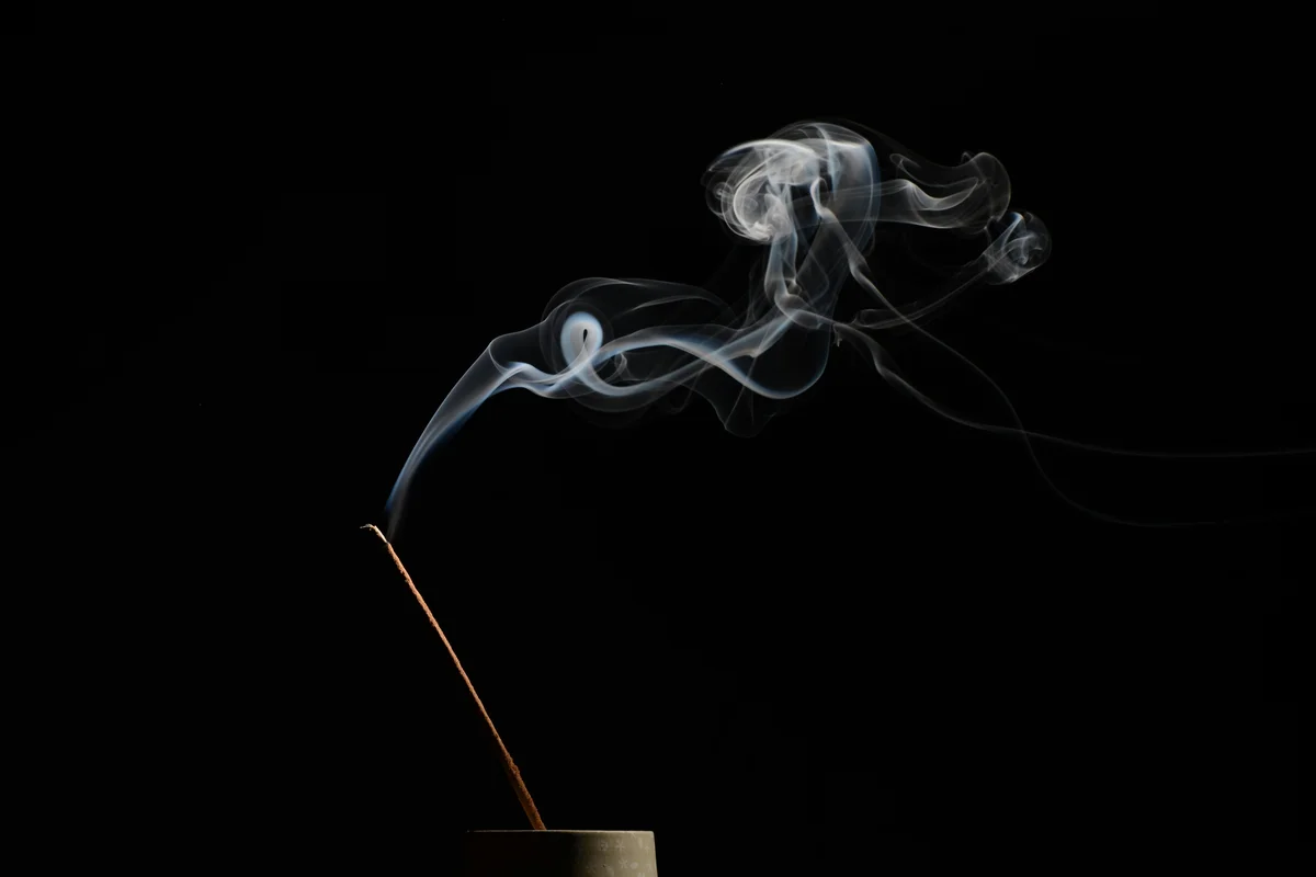 A single lit incense stick in a small stone holder against a pure black background, with a trail of white smoke curling and billowing upward in an intricate flowing pattern