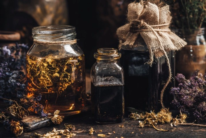 Three glass jars of different sizes on a dark wooden surface (one sealed with golden honey and dried herbs, one containing dark liquid with salt, one tied with linen twine) surrounded by scattered dried botanicals and a lit beeswax candle in warm candlelight