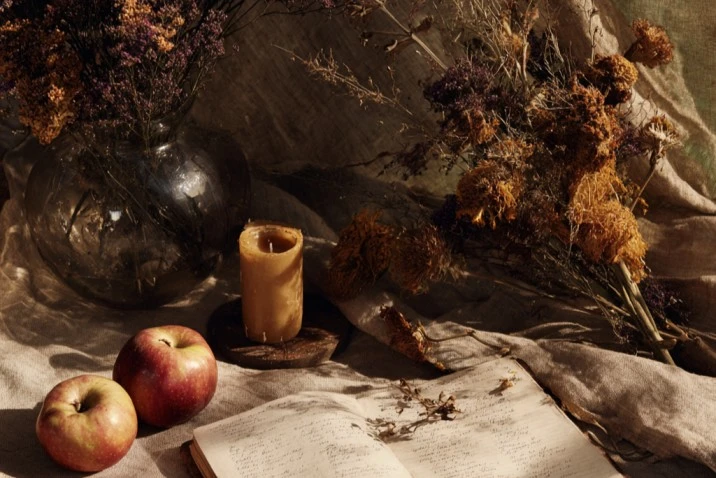 Apples cut horizontally to show their seed pentagrams beside an open journal with handwritten autumn intentions, dried marigolds, and a single beeswax candle on autumn-toned linen in soft amber light