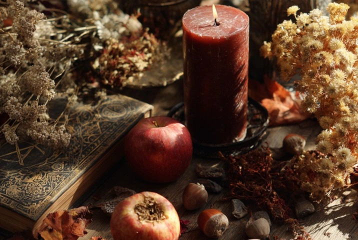 A Mabon altar on a wooden surface with two red apples, a cut apple showing its pentagram of seeds, dried autumn leaves in rust and gold, a deep red candle, a handful of acorns and hazelnuts, and a sprig of dried chrysanthemum in warm afternoon light