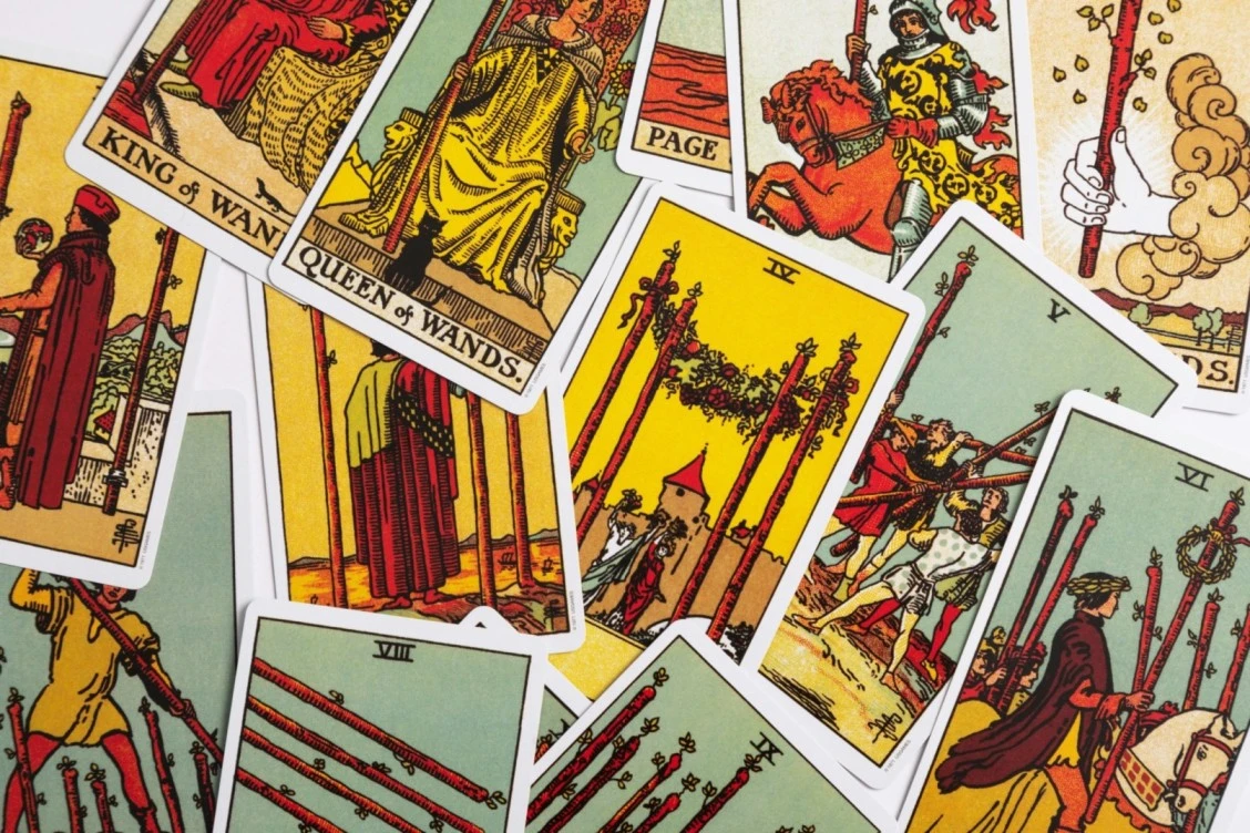 The four Aces of a tarot deck arranged in a row on dark linen — Wands, Cups, Swords, Pentacles — with a small white candle burning beside them in low warm candlelight