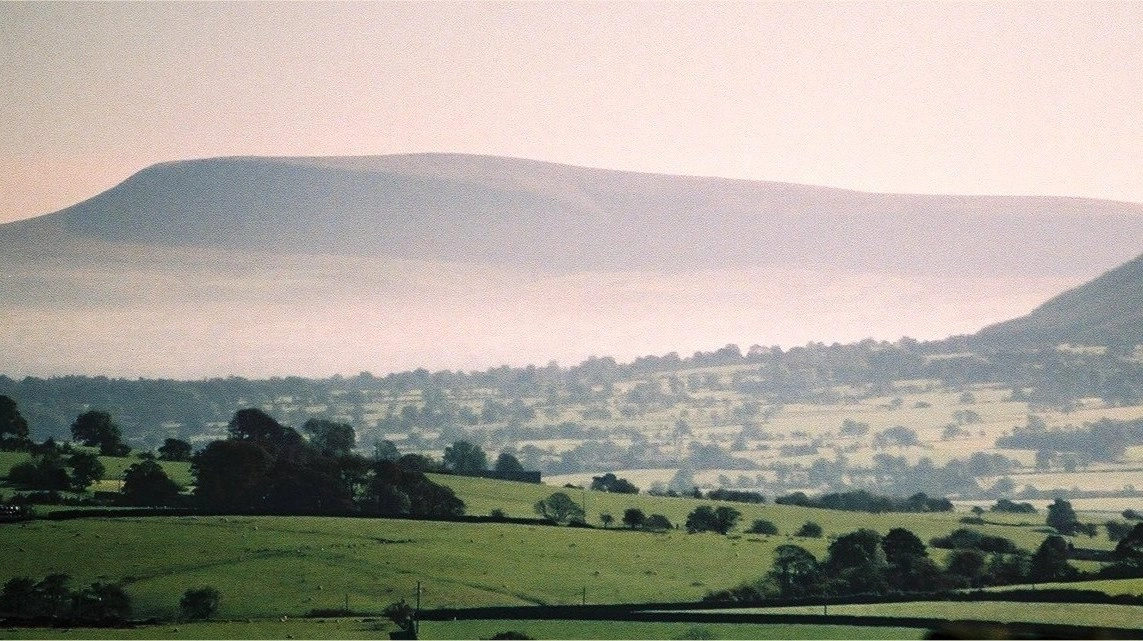 Pendle Hill rising in the distance above rolling Lancashire fields, with low mist drifting across hedgerows in the foreground at dusk