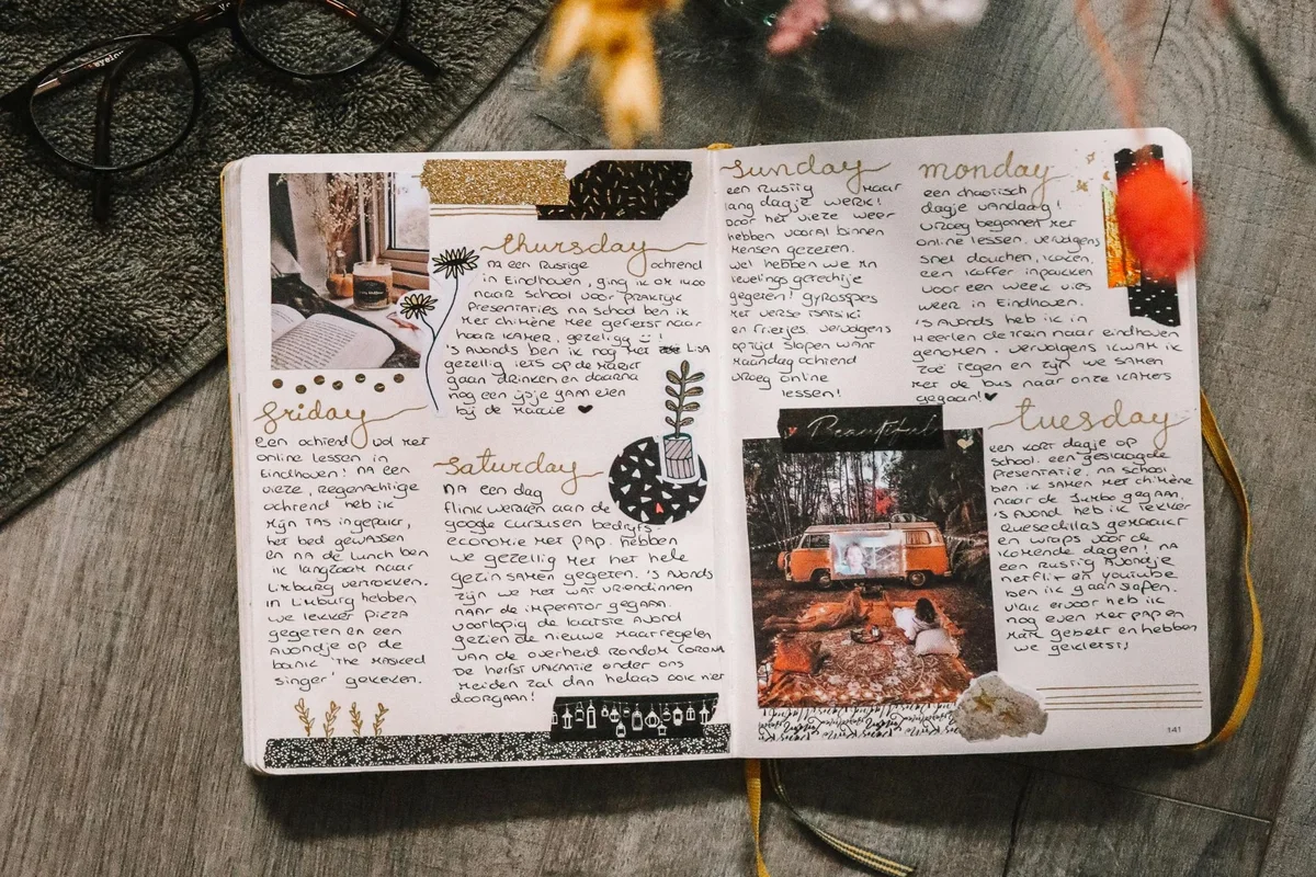 An open journal spread across two pages filled with handwritten daily entries for Thursday through Tuesday, decorated with washi tape, small photographs, botanical doodles, and gold accents, resting on a wooden surface beside reading glasses and a raw crystal