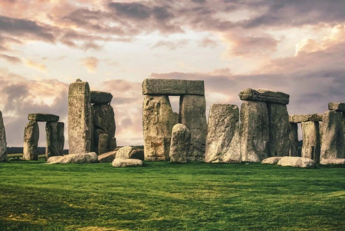 Stonehenge in the Wiltshire landscape at dawn, the great sarsen trilithons silhouetted against a cool pink solstice sky, with morning mist drifting low across the chalk downland