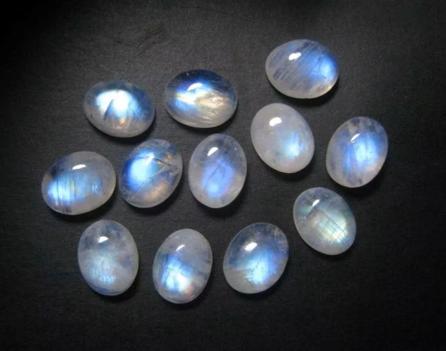 A scattered collection of oval cabochon moonstones showing strong blue adularescent flash, on a dark surface