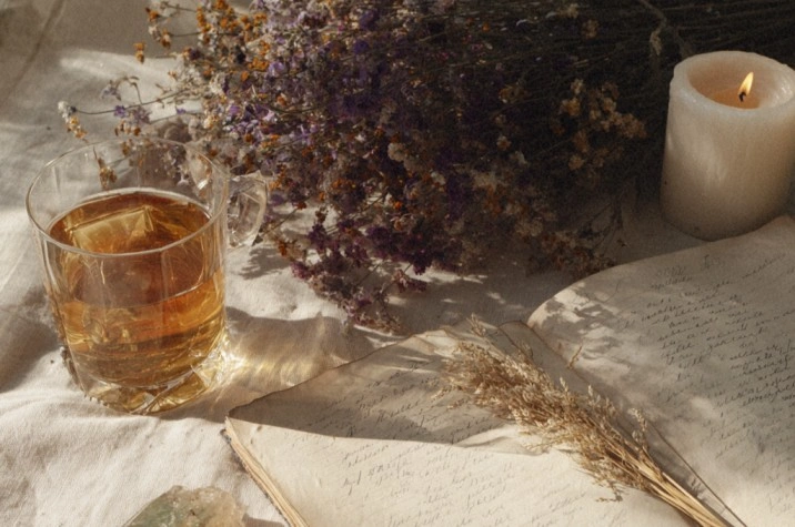 A late-summer flat lay with a glass of chamomile tea, an open notebook with handwritten lists, a sprig of dried lavender, a piece of peridot, wheat stalks, and a lit white candle on cream linen in clear morning light