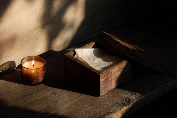 A folded slip of paper inscribed with a single handwritten name resting inside an open wooden box beside a lit beeswax candle on a dark surface in low warm light