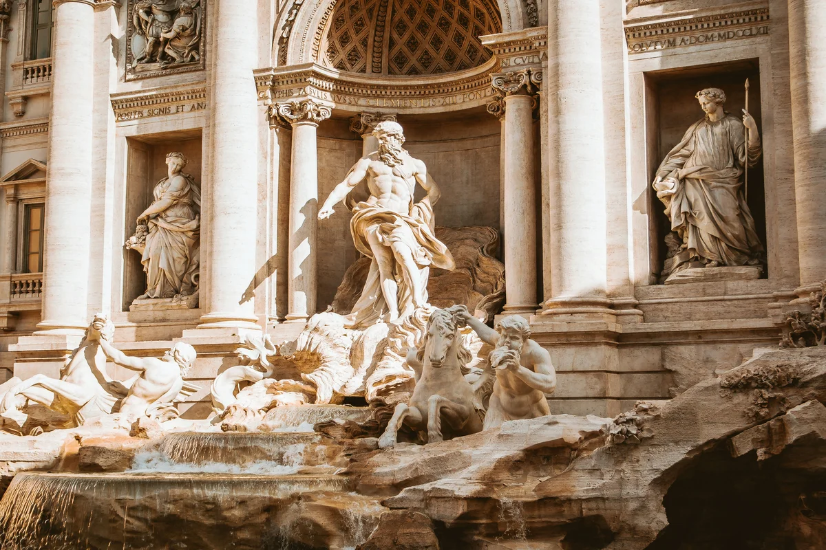 The Trevi Fountain in Rome, showing the central figure of Neptune commanding the waters from a triumphal arch facade, flanked by allegorical statues, seahorses, and tritons in warm golden light