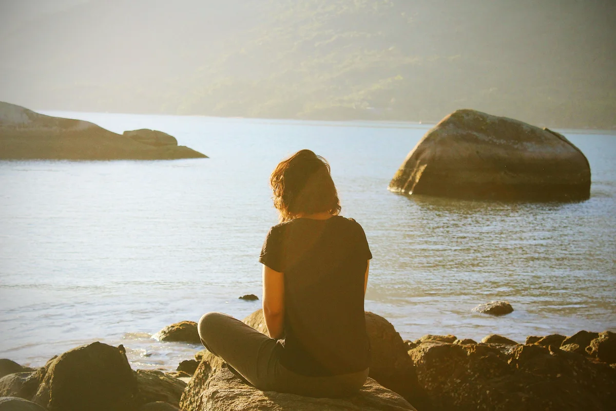 A person with short auburn hair sitting cross-legged on coastal rocks, seen from behind, gazing out over still water toward large boulders and misty hills in warm golden light