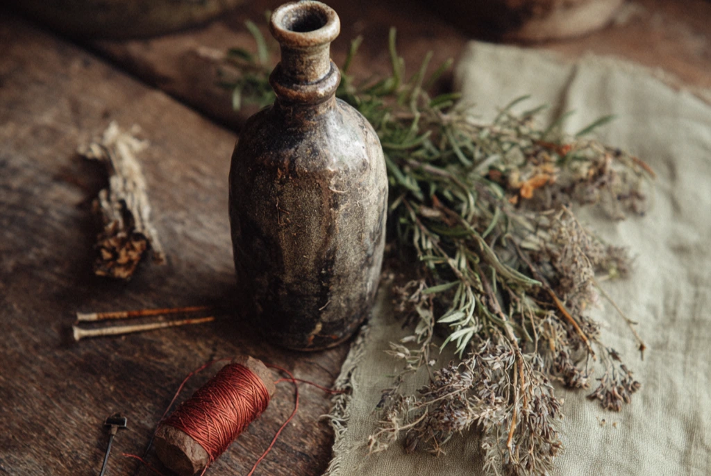A small earthenware bottle on a wooden surface beside iron pins, a sprig of rosemary, dried protective herbs, and a length of red thread, photographed in low warm candlelight against dark linen