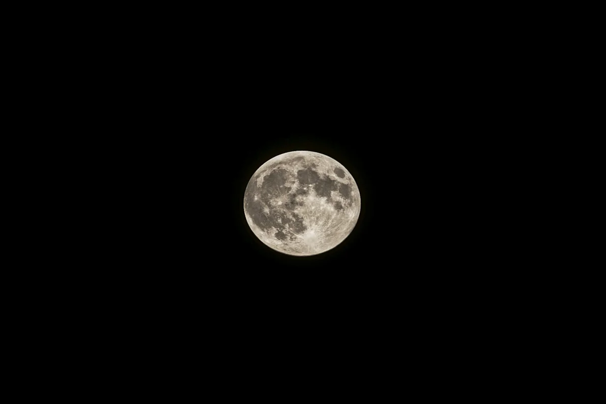 A full moon photographed against a pure black night sky, showing surface detail including craters and maria in shades of grey and warm white